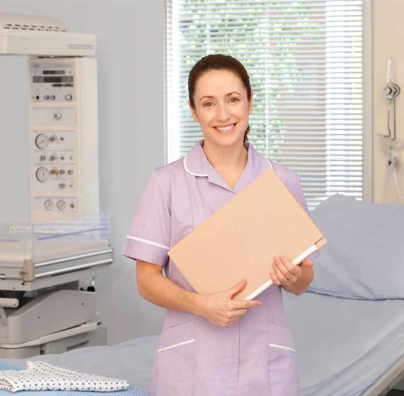 Uśmiechnięta położna w rózowym fartuchu, trzymająca dokumentację medyczną, stojąca w gabinecie lekarskim i patrząca w obiektyw.