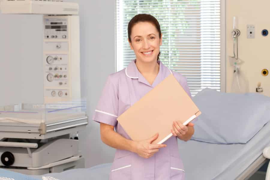 Uśmiechnięta położna w rózowym fartuchu, trzymająca dokumentację medyczną, stojąca w gabinecie lekarskim i patrząca w obiektyw.